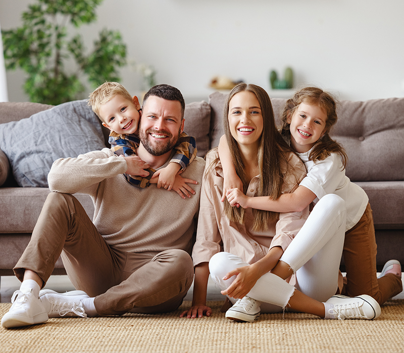 Happy family of four sitting together on floor in front of couch, smiling at camera.